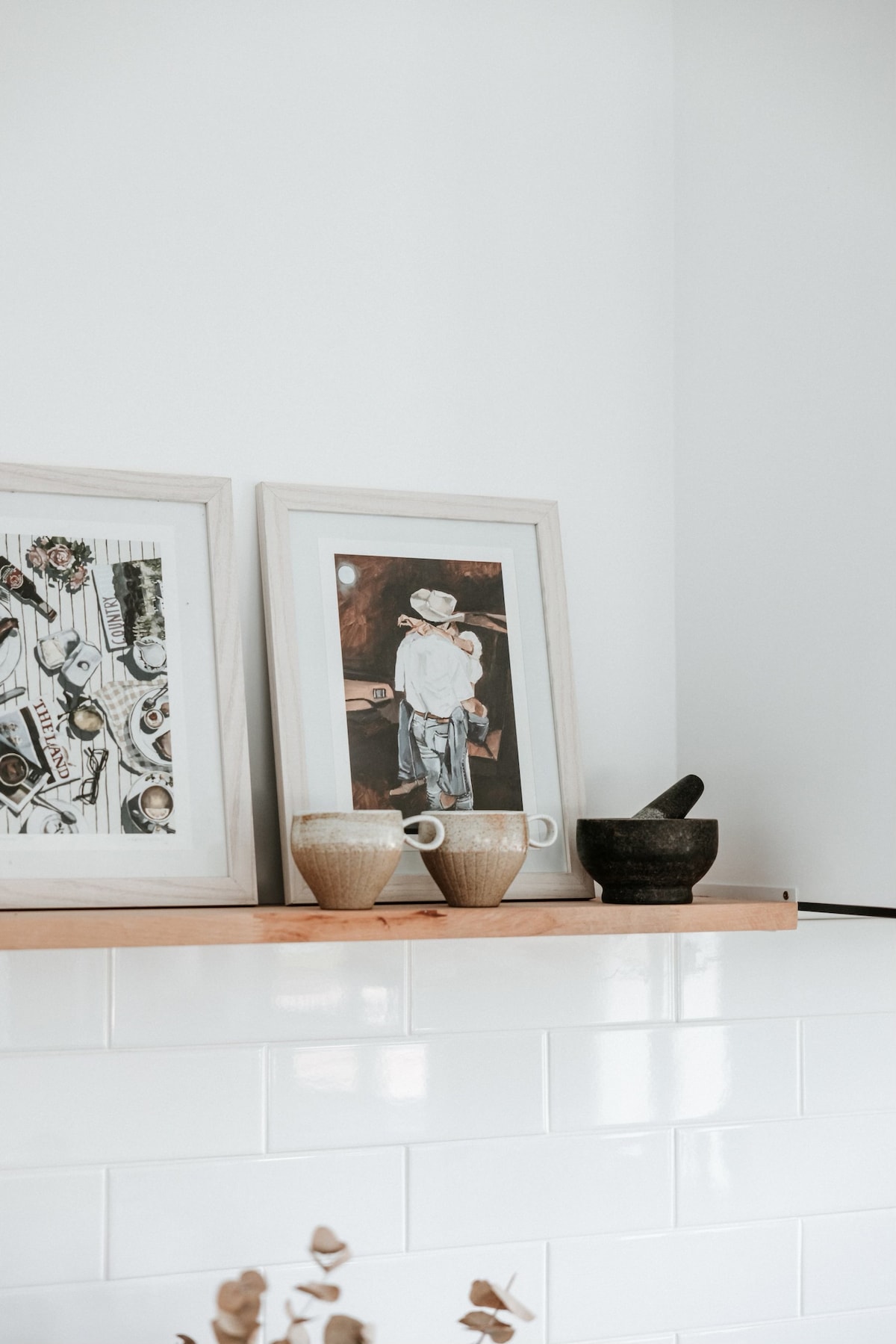 A wooden shelf displays two framed artworks alongside ceramic and stone vessels. The light-colored frames complement the white wall, while the textures of the pottery add an organic touch to the subtle decor.