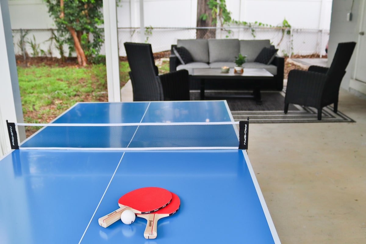 An outdoor recreational area features a bright blue ping pong table, with a paddle and ball placed on its surface. In the background, a seating arrangement includes a gray sofa and chairs, surrounded by greenery and a light rug.