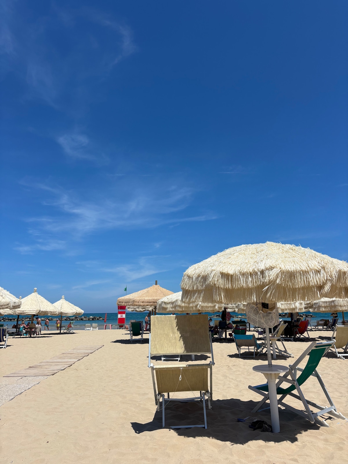 A beach scene is presented with numerous straw umbrellas providing shade over sun loungers. The golden sand stretches towards a clear blue sky, with groups of people enjoying the beach in the background. The gentle ripples of the calm sea are visible in the distance.