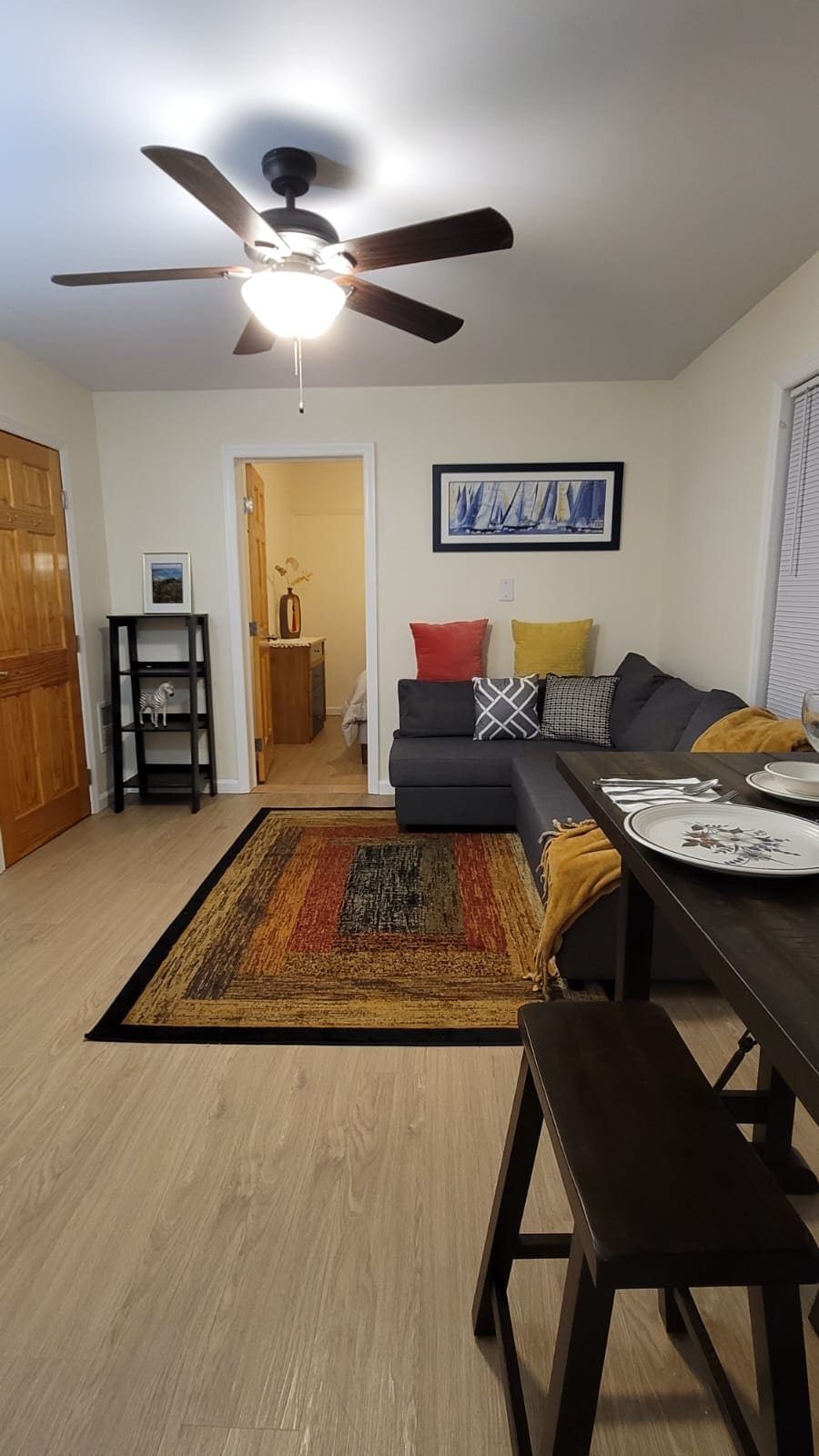 A cozy sitting area is highlighted by a spacious gray sofa adorned with colorful throw pillows. A dining table with stools is positioned nearby, while a ceiling fan provides ventilation. Natural light enters through a window, illuminating the space and wooden accents.