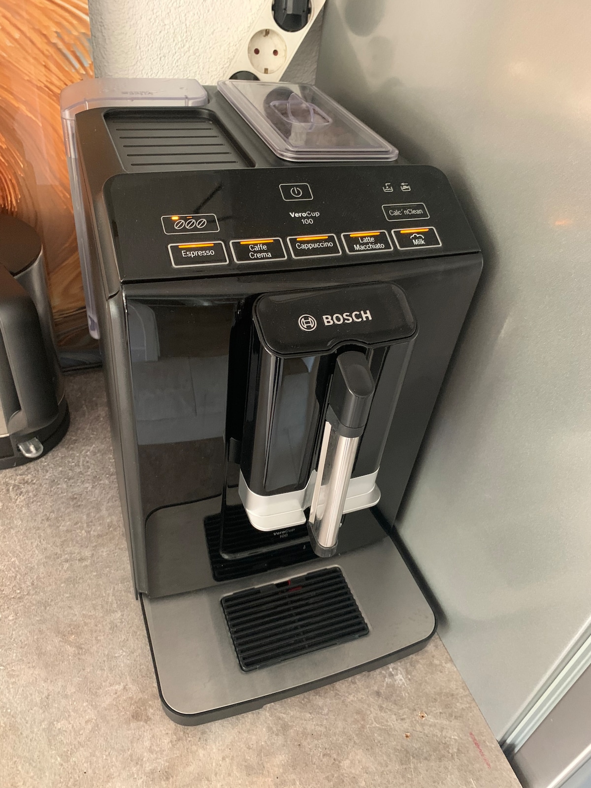 A Bosch coffee machine is positioned on a countertop, featuring multiple buttons for various brewing options. The sleek design includes a milk frother attachment and a clear container for holding coffee beans. The machine's black exterior complements the surrounding kitchen decor.