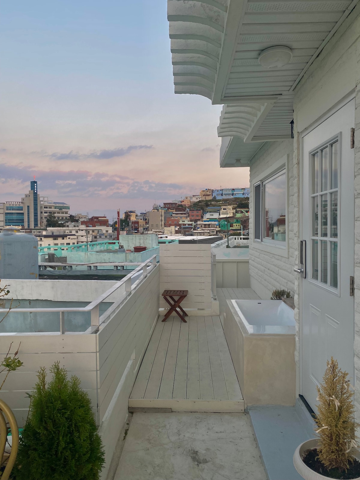 A serene balcony space is presented, featuring a view of the surrounding cityscape at dusk. A small stool and a bathtub are positioned on the wooden deck, which is bordered by white walls. Soft colors of the sunset reflect off the buildings in the background.