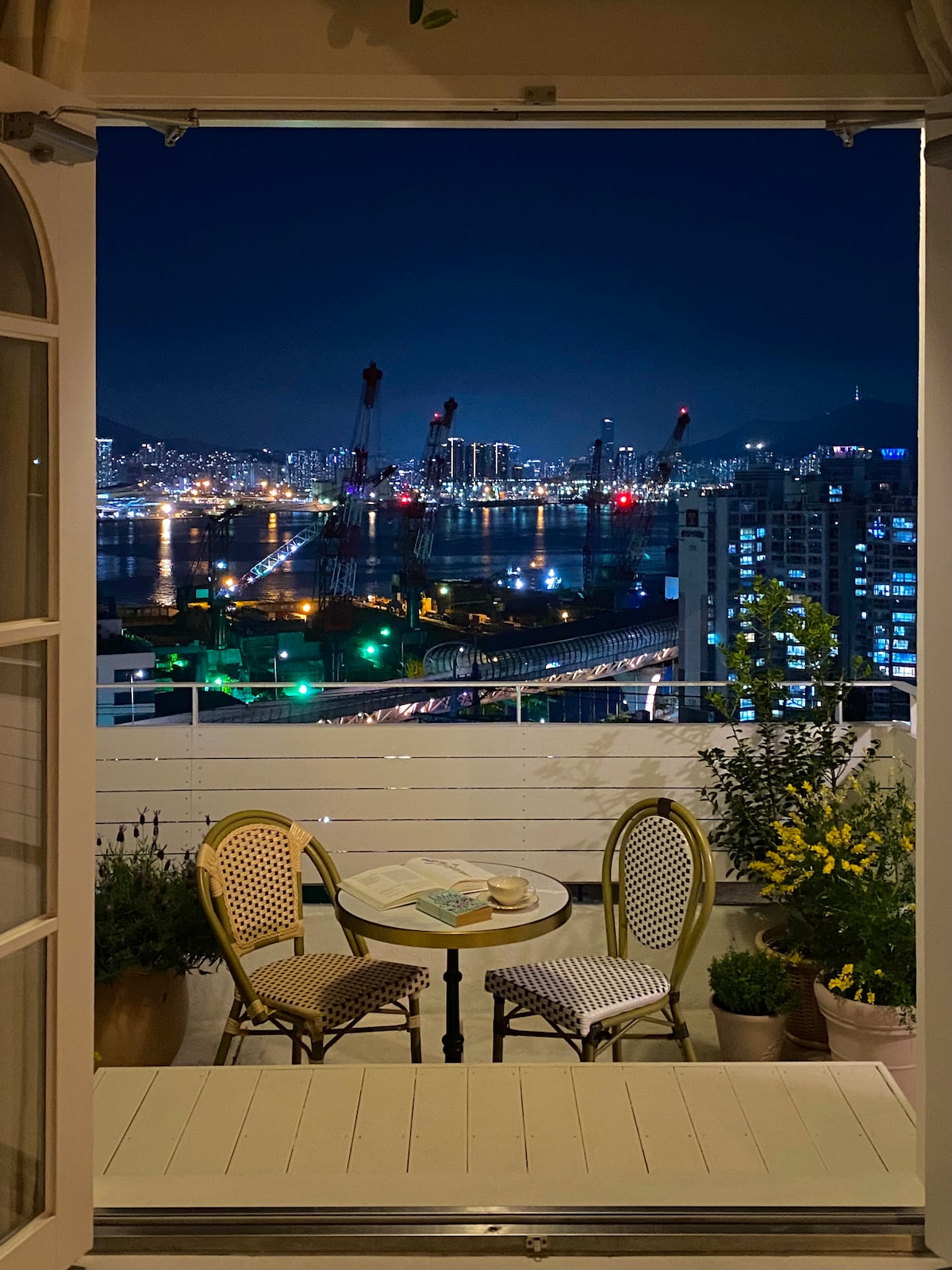 A small balcony is featured with two round chairs and a table, set against a backdrop of a night view over the city and harbor. Soft lighting illuminates the scene, while cranes and buildings outline the horizon, creating a serene atmosphere.