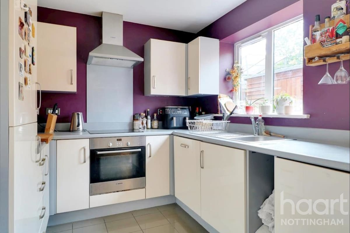 A well-equipped kitchen features white cabinetry paired with a deep purple accent wall. Stainless steel appliances, including an oven and a hood, are visible. A sink and countertop provide ample workspace, and natural light enters through a window adorned with potted plants.