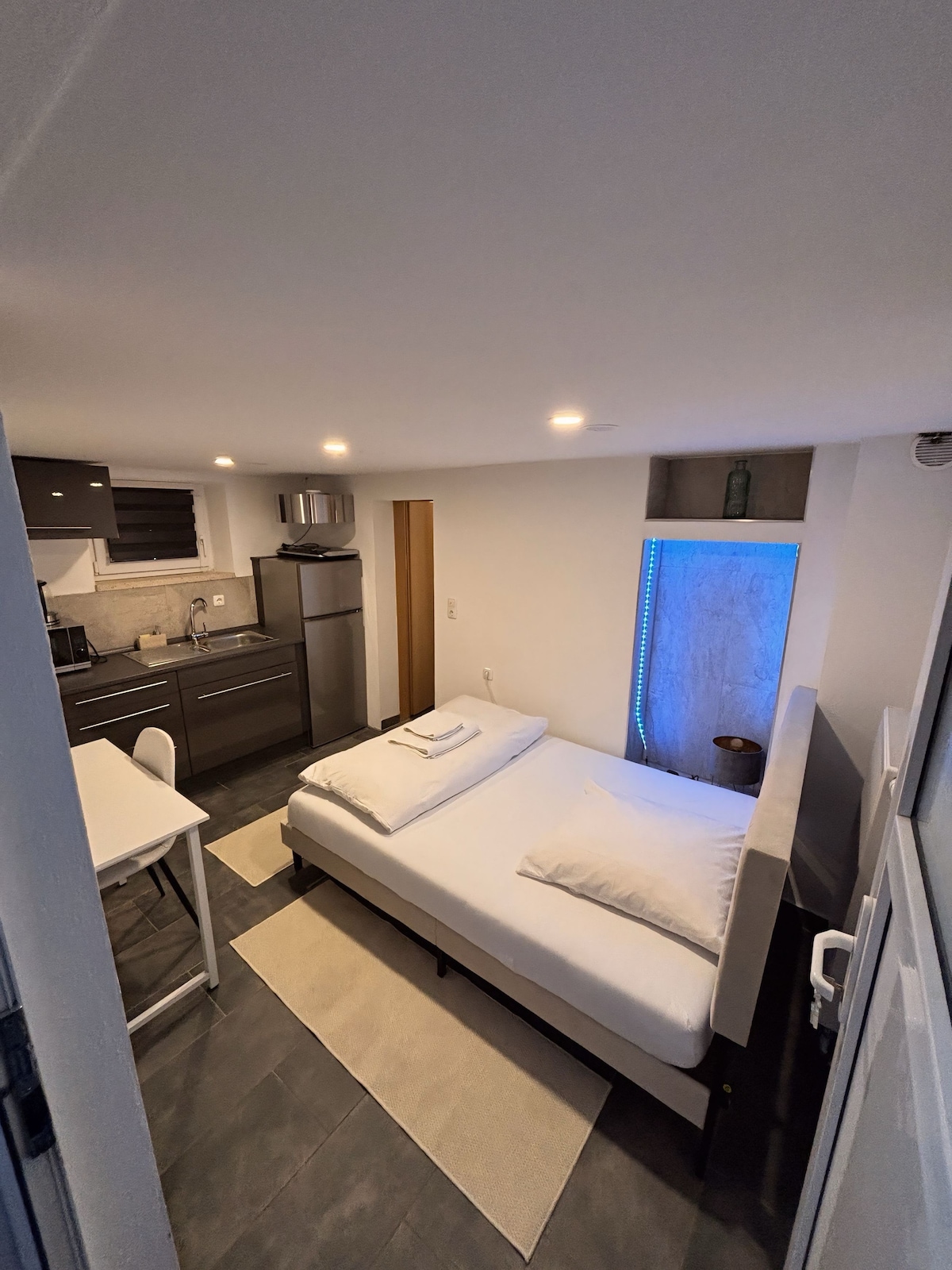 An open sleeping and kitchen area features a spacious bed with neatly arranged linens. A compact kitchen is visible, equipped with modern appliances. Soft LED lights illuminate the space, complemented by a light-colored area rug that adds comfort to the floor.