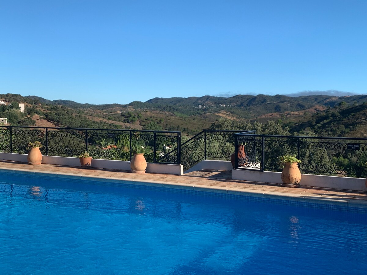 A tranquil private pool reflects the clear blue sky, surrounded by lush greenery and rustic terracotta pots. The mountainous landscape provides a serene backdrop, enhancing the peaceful ambiance of the outdoor space.