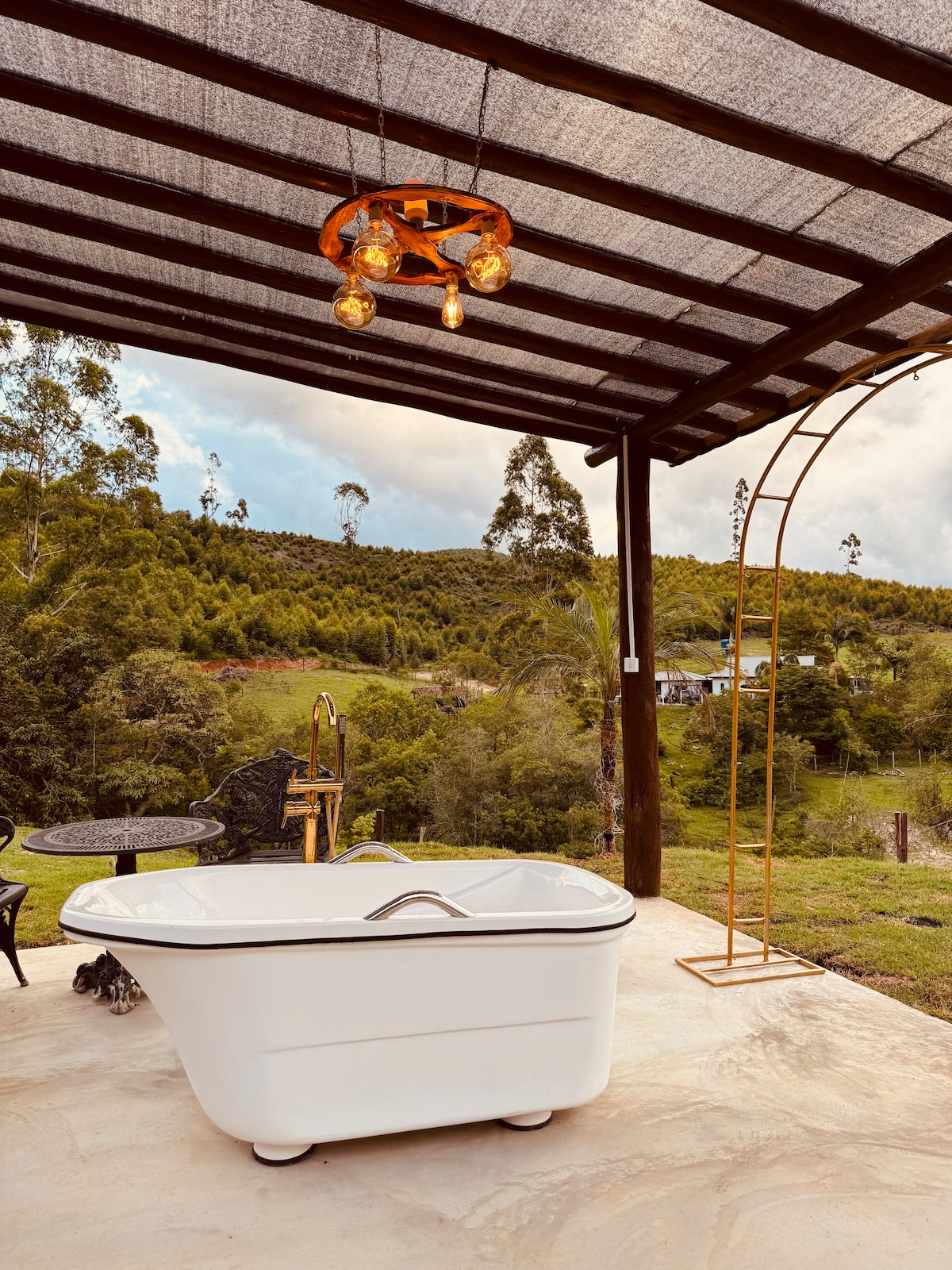 An outdoor area features a freestanding white bathtub under a canopy with rustic lighting. A wrought iron table and chairs are arranged nearby, surrounded by lush greenery and rolling hills in the background. The space offers a serene spot for relaxation.