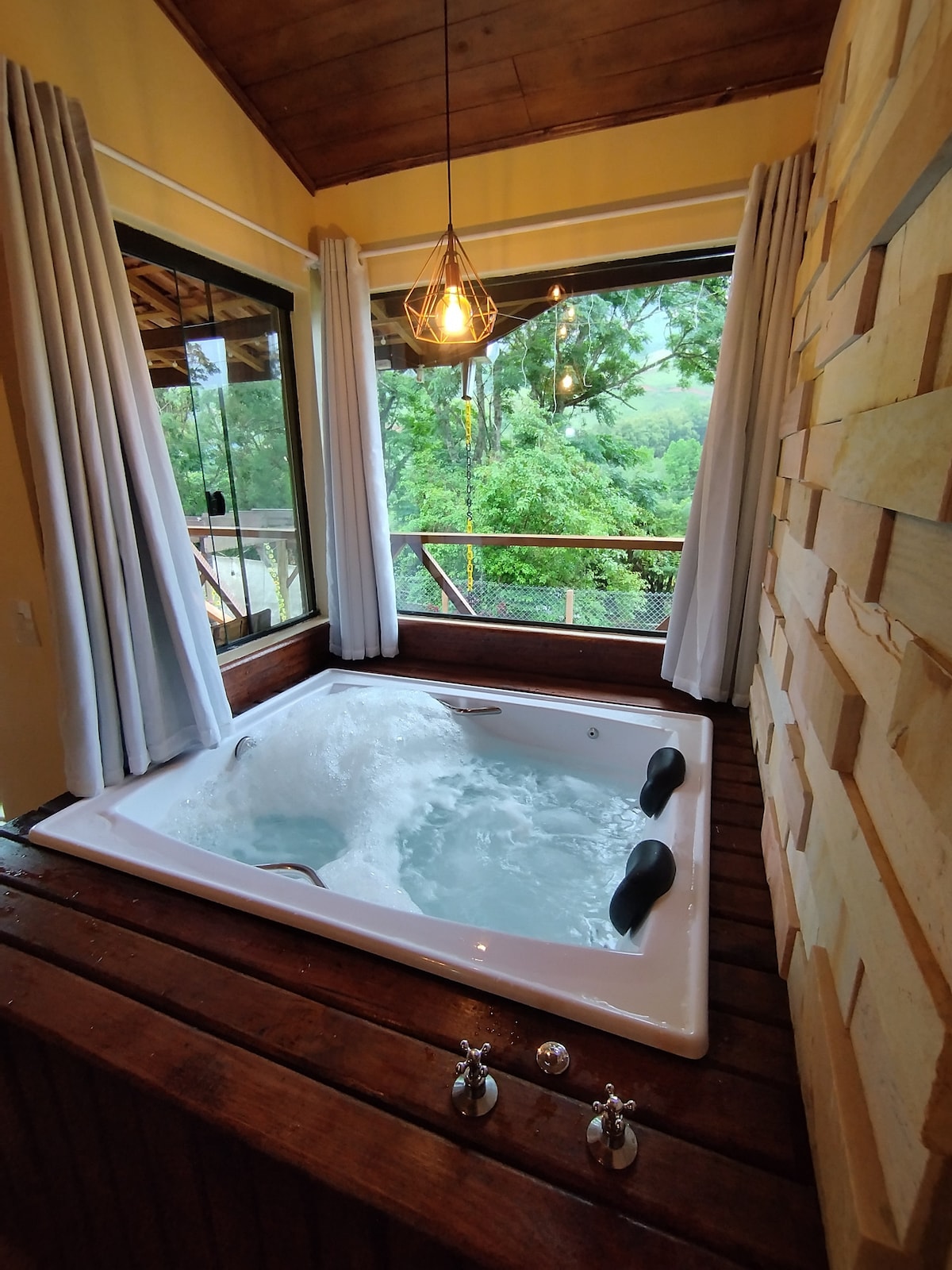 A spacious hydro-massage bathtub is positioned near large windows, allowing natural light to illuminate the tranquil space. The surrounding wooden deck features an organic aesthetic with natural wood paneling, enhancing the overall serene atmosphere.