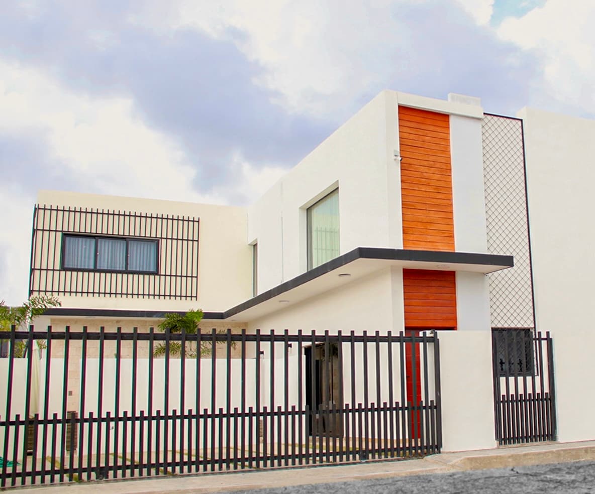 The villa's modern exterior is characterized by a combination of sleek white walls and warm wooden accents. A secure black gate encloses the property, with neatly manicured greenery visible through the openings. Large windows enhance the contemporary design, allowing natural light to flood the interior.