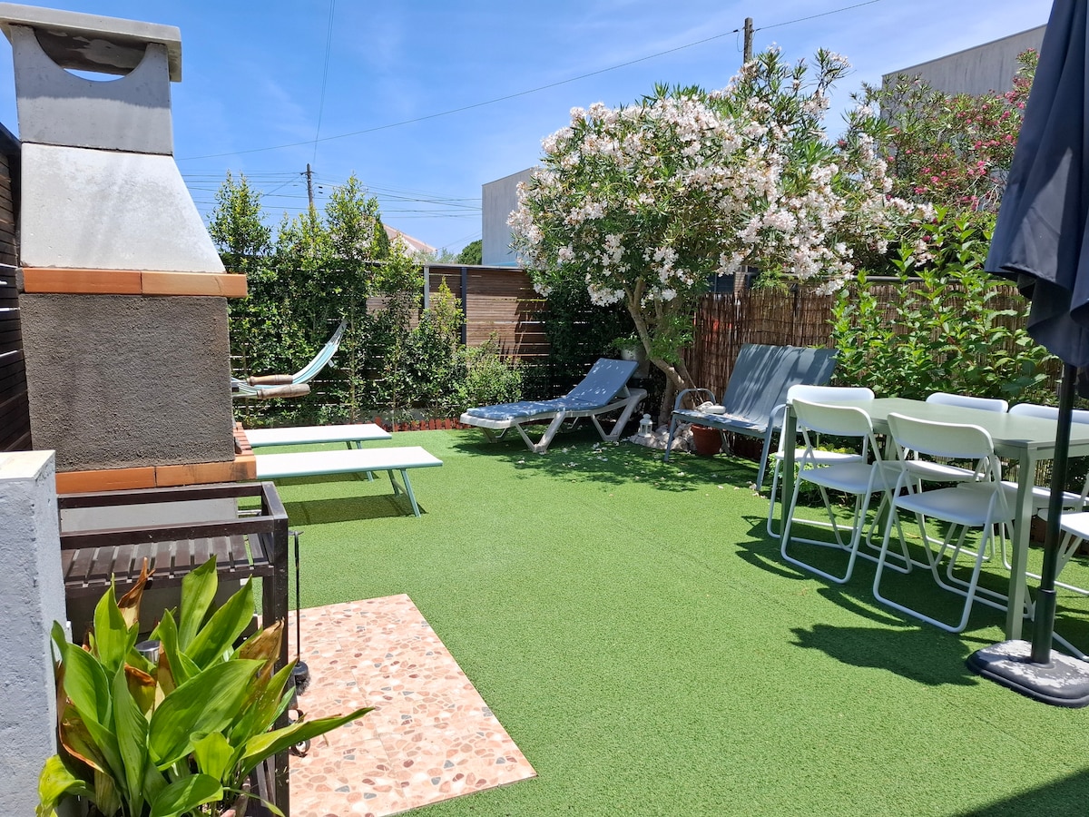 A landscaped outdoor space includes a green lawn area, sun loungers, and a dining table with chairs. A stone barbecue is positioned nearby, surrounded by vibrant foliage and flowering bushes, creating a serene environment for relaxation and social gatherings.