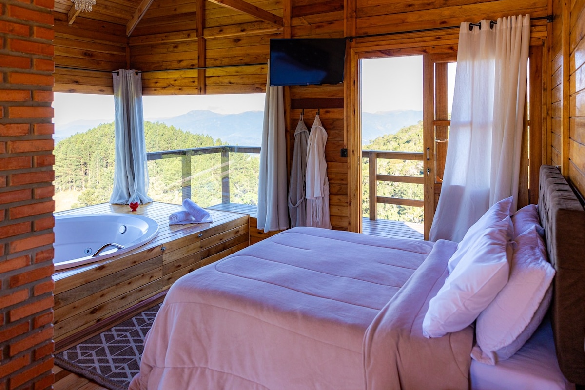 A cozy room features a spacious bed with soft linens and a bath area with a jetted tub beside large windows. Natural wood accents enhance a calm ambiance, and a balcony offers a scenic view of the surrounding hills. A television is mounted on the wall.