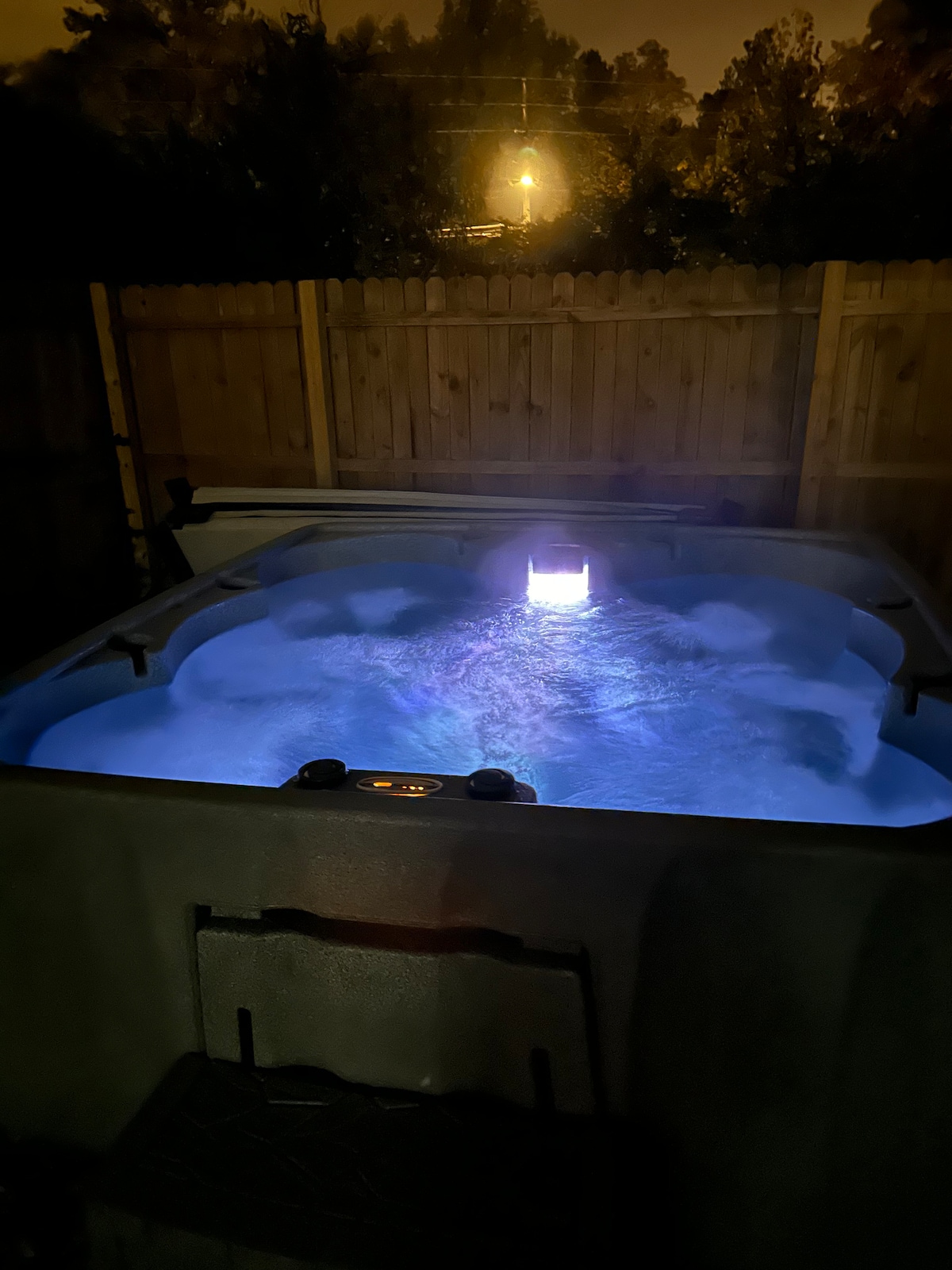 A hot tub is seen illuminated by soft lighting, creating a serene ambiance. The bubbling water suggests a soothing experience. A wooden fence surrounds the area, providing privacy, while ambient light is visible in the background.