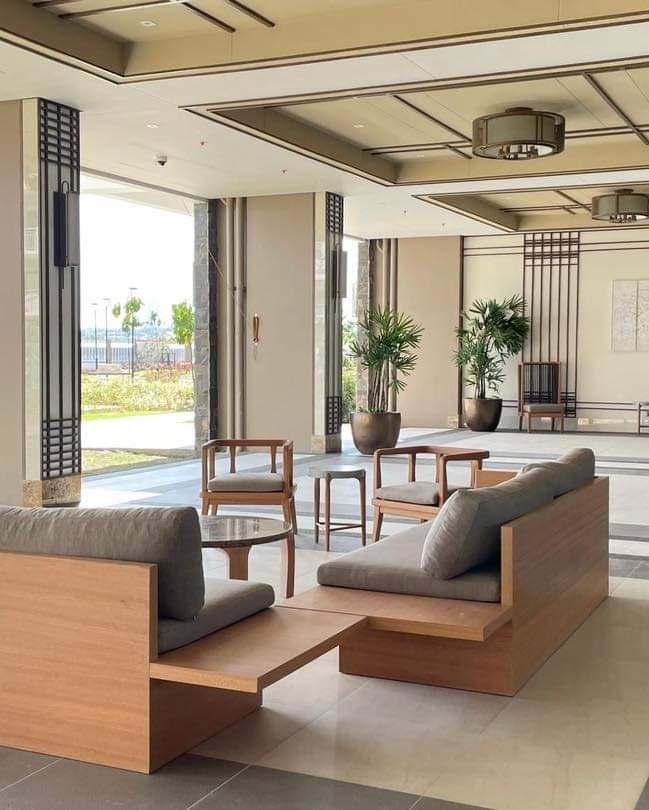 A modern lobby area is shown with a comfortable wooden seating arrangement, featuring a low bench and a couple of single chairs. Lush indoor plants are positioned nearby, contributing to a refreshing ambiance. Large windows allow natural light to fill the space.