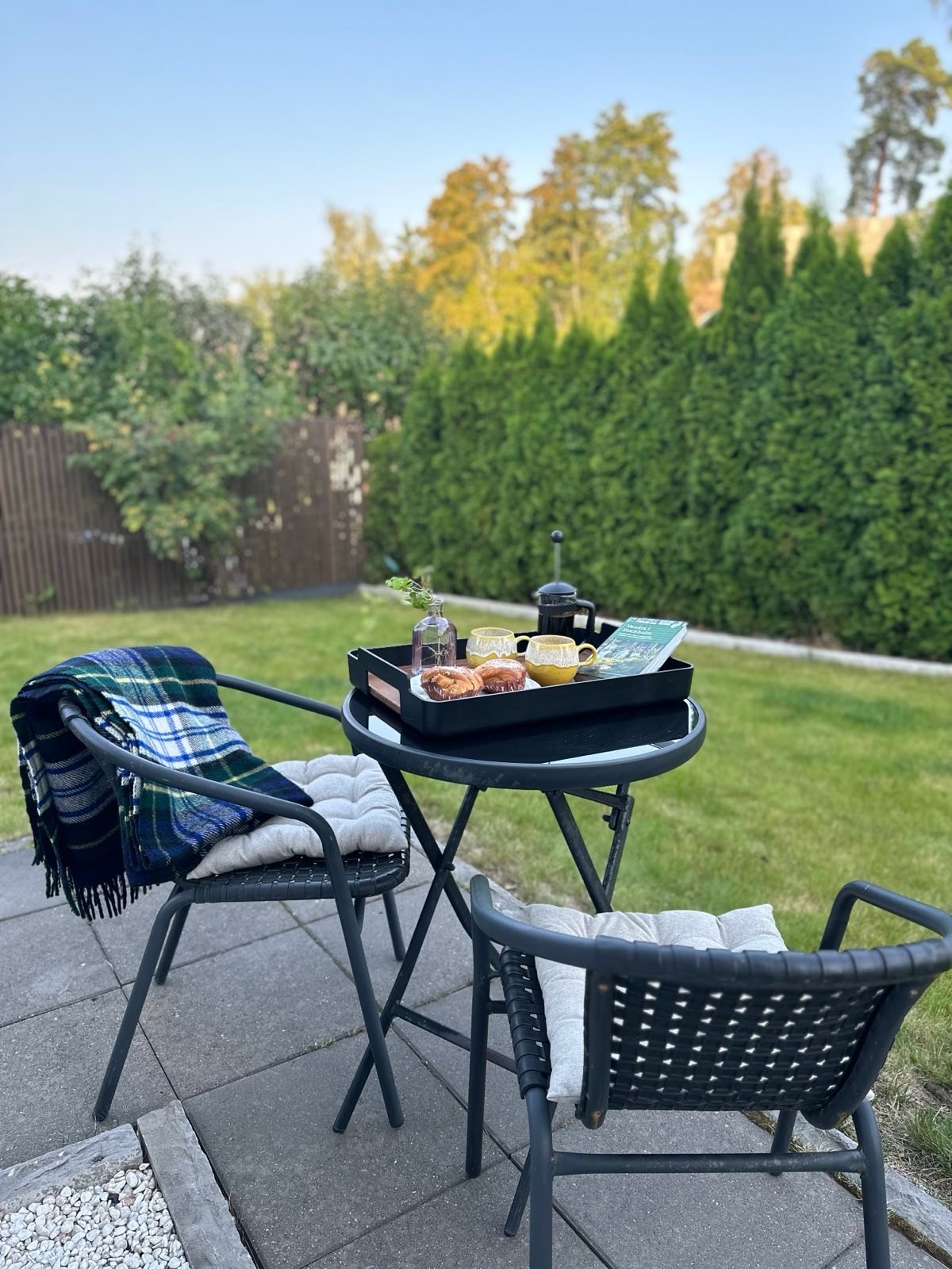 A small outdoor seating area is set with a round table and two chairs. A tray holds a coffee pot, cups, pastries, and a book. A cozy plaid blanket drapes over one chair, surrounded by lush greenery and a clear blue sky.