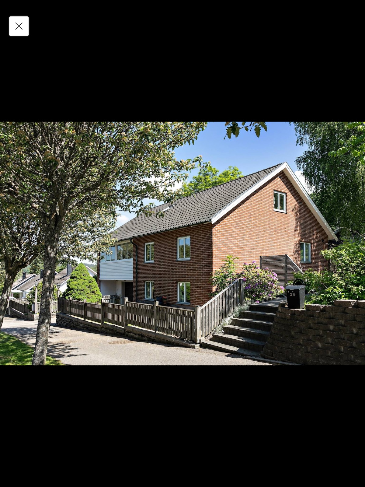 A well-maintained two-story brick house is framed by lush trees and shrubs. A wooden fence lines the front, with a pathway leading up to the entrance steps, enhancing the property’s welcoming appearance. Sunlight filters through the foliage, creating a pleasant atmosphere.