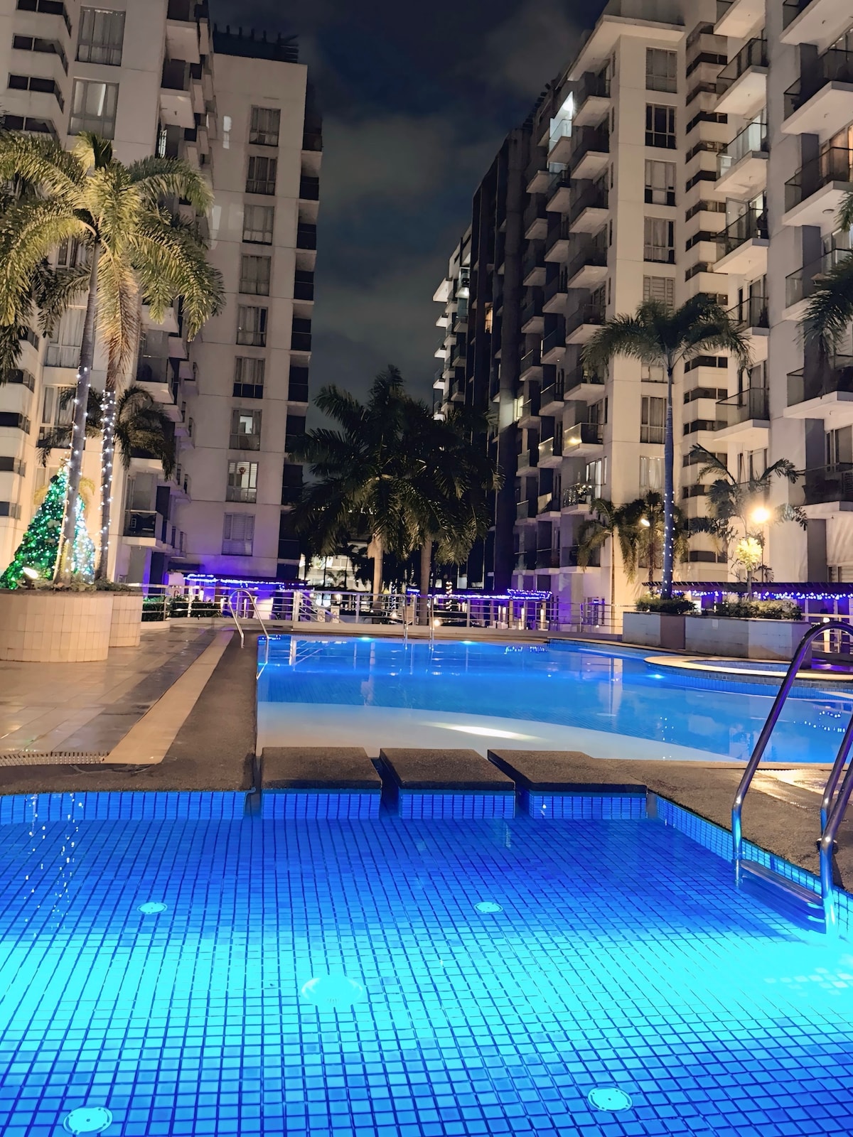 The image showcases a serene pool area illuminated by soft blue light, surrounded by modern buildings. Palm trees line the poolside, enhancing the tranquil setting. The pool features steps leading into the water, inviting guests to relax and unwind in this refreshing space.