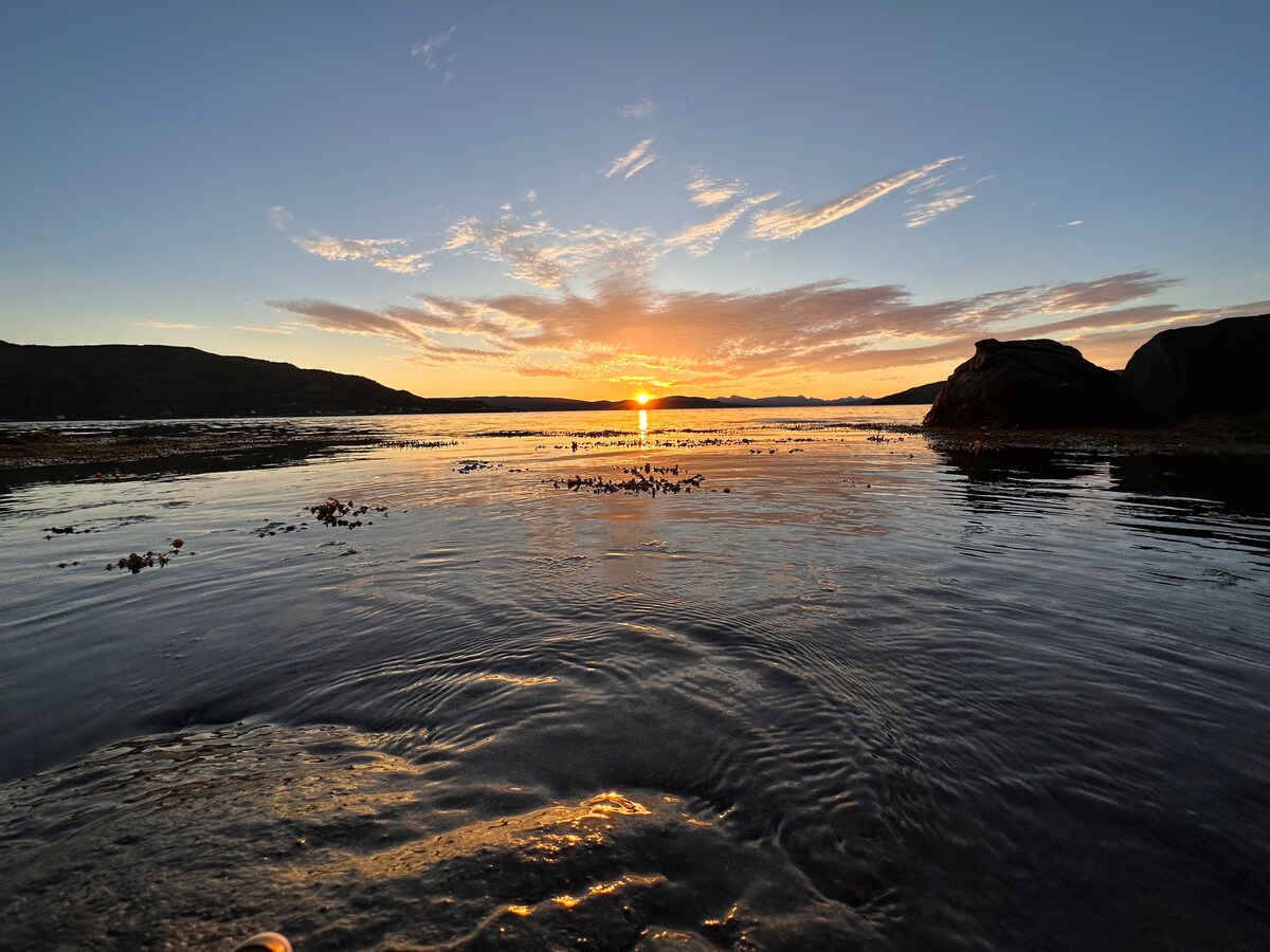 The image captures a serene sunset over calm waters, with soft waves creating gentle ripples. Silhouettes of hills can be seen in the background, while clouds are softly illuminated by the setting sun, adding warmth to the evening sky.