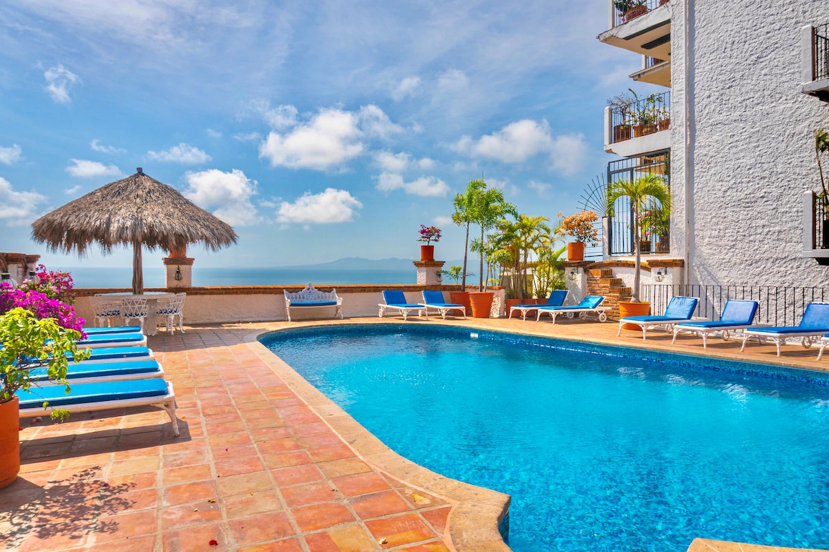 A bright and inviting outdoor space features a large swimming pool surrounded by lounge chairs. A thatched-roof palapa offers shaded seating, while potted plants and vibrant flowers add color to the scene. The bay is visible in the background, enhancing the tranquil atmosphere.