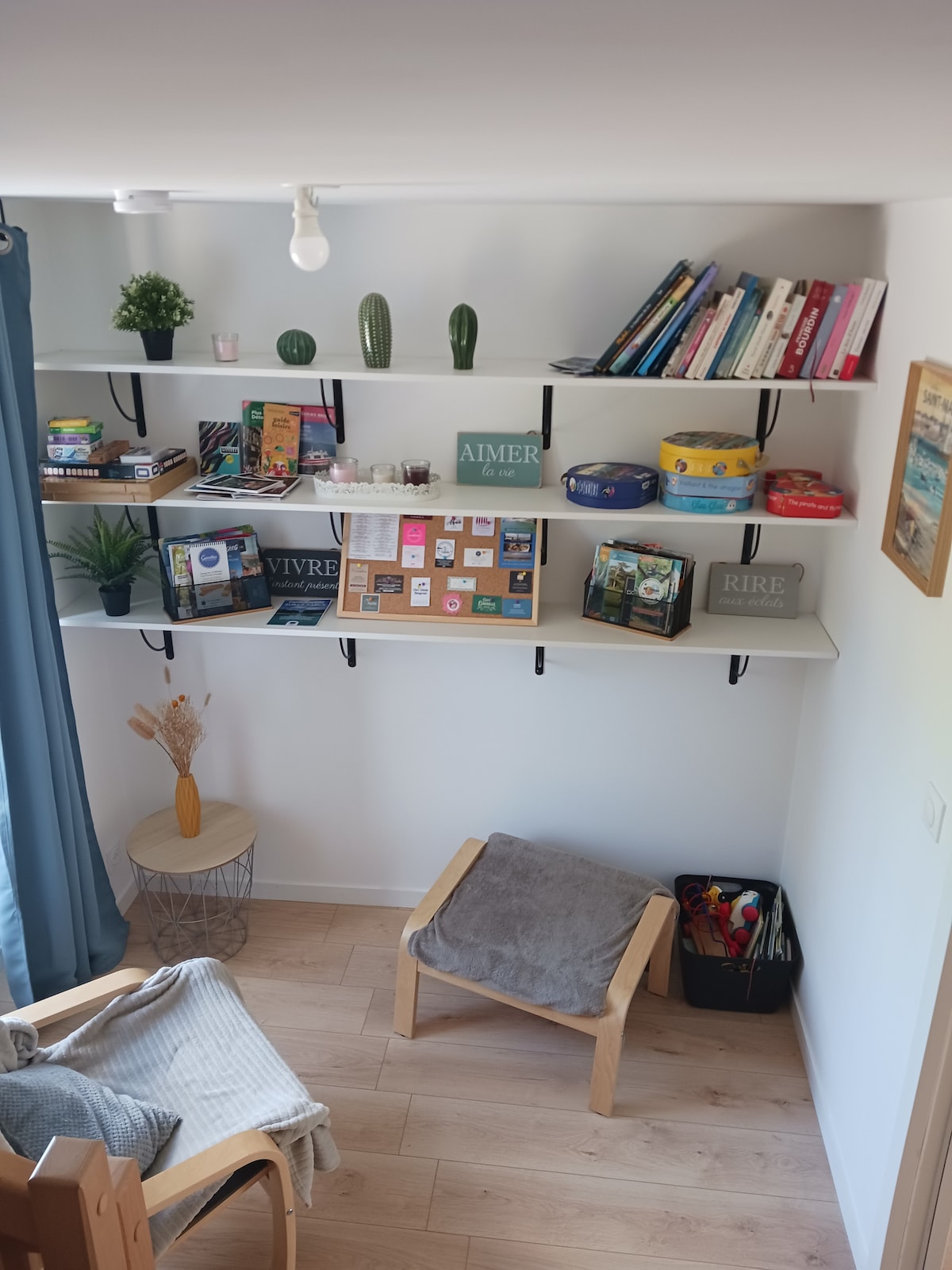 A cozy reading nook features a wooden chair with a soft gray throw and a small side table. Shelving units display an assortment of books, games, and decorative items, including indoor plants, creating a warm and inviting space for relaxation and leisure.
