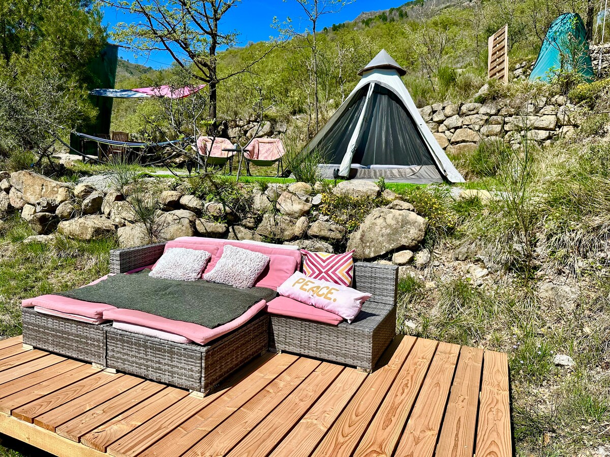 An outdoor seating area is placed on a wooden deck, featuring a cushioned sofa adorned with soft pillows. In the background, a tipi structure is visible amidst a lush, green landscape, enhanced by natural stone formations and gentle sunlight.