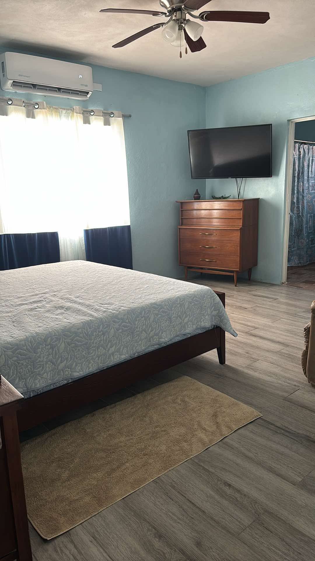 A spacious sleeping area is furnished with a king-size bed covered by a light-colored blanket. Natural light filters through large windows with dark curtains. A mounted television is placed across from the bed, and a wooden dresser occupies one corner of the room. A bathroom is visible in the background.