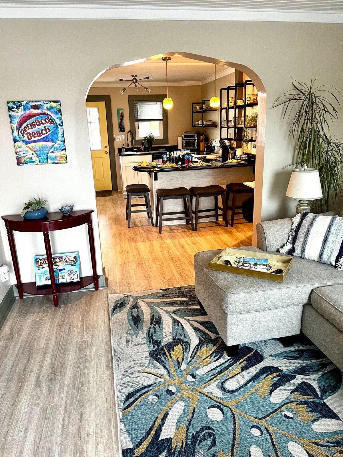 The cozy living area is adorned with a patterned area rug and a gray sectional sofa. An archway leads to a modern kitchen featuring a breakfast bar with stools. The walls are painted in warm tones, complemented by vibrant wall art, creating an inviting space.