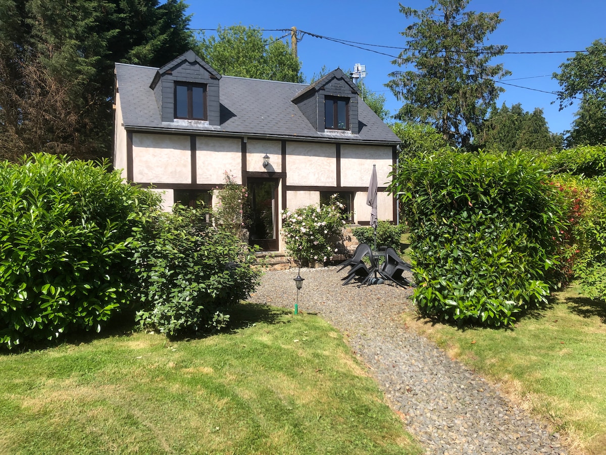 A charming two-bedroom cottage sits surrounded by lush greenery, set against a clear blue sky. The entrance is enhanced by a stone pathway, with a patio area featuring outdoor seating. The garden is well-maintained, inviting a sense of tranquility and privacy.