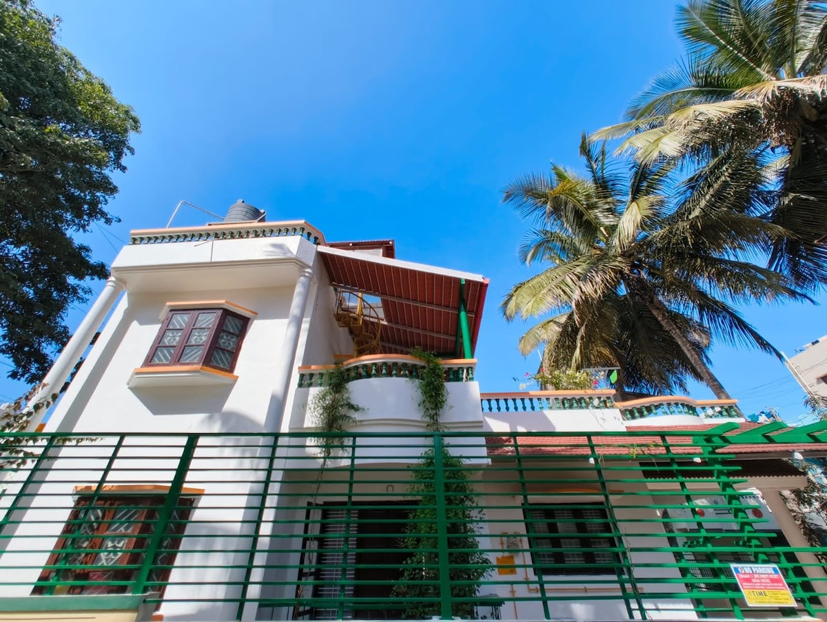 A two-story home with a clean white facade is framed by lush green plants and palm trees. The balcony is visible on the upper level, surrounded by decorative railings. Bright blue skies create a vibrant backdrop, enhancing the welcoming appearance of the exterior.