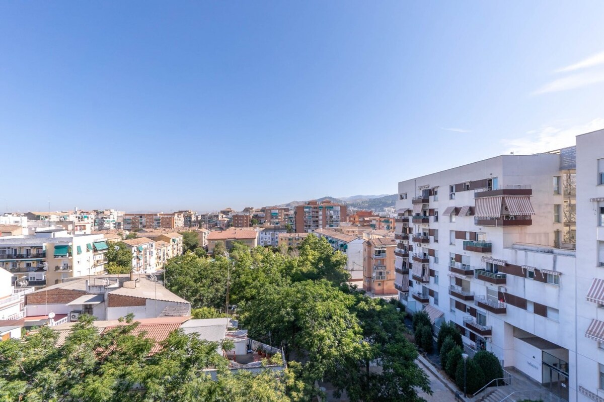 A panoramic view of a residential neighborhood showcases a mix of buildings and greenery. The scene includes rooftops and trees, with distant mountains visible under a clear blue sky, providing a sense of tranquility and openness.