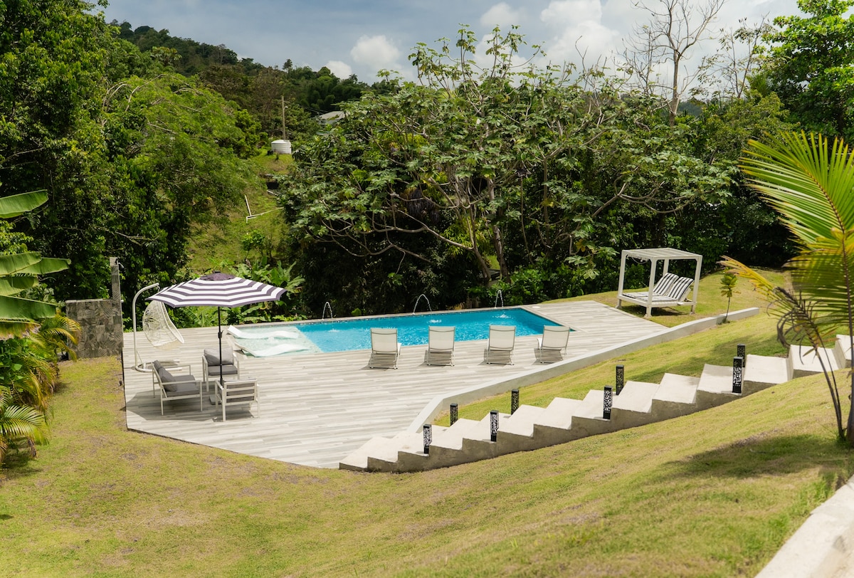 A serene pool area is surrounded by lush greenery, featuring a rectangular swimming pool with lounge chairs positioned around it. A striped umbrella offers shade, while a swinging bench adds to the outdoor space. The landscape gently slopes down towards the pool deck.