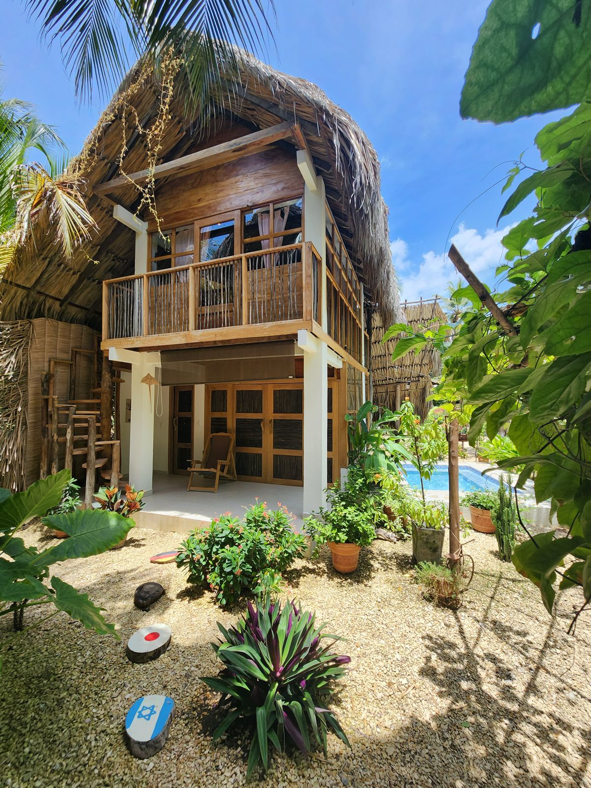 A charming two-story building features a thatched roof and wooden framework, surrounded by lush greenery. Potted plants and colorful foliage enhance the vibrant garden area, while a glimpse of a swimming pool is visible in the background under the clear blue sky.
