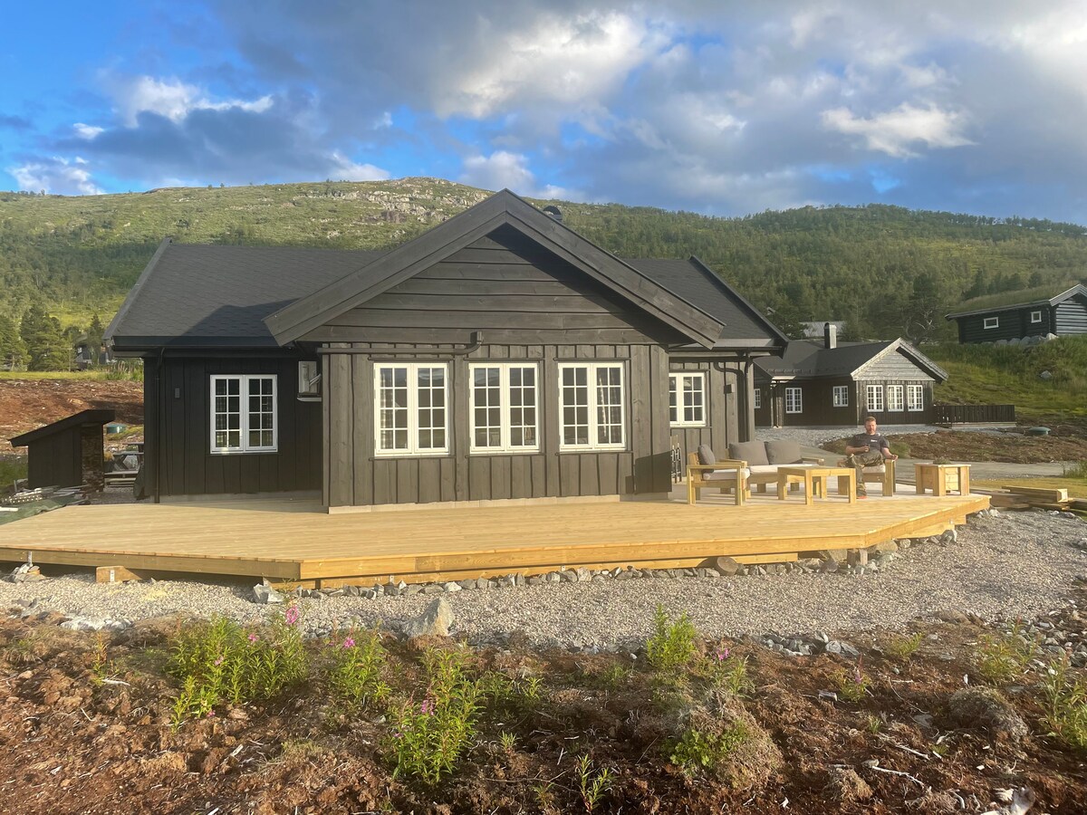 A charming cabin is set against a backdrop of rolling hills and trees. A spacious wooden deck extends from the front, furnished with a table and benches. Multiple windows provide natural light, while the dark exterior contrasts with the light-colored trim.