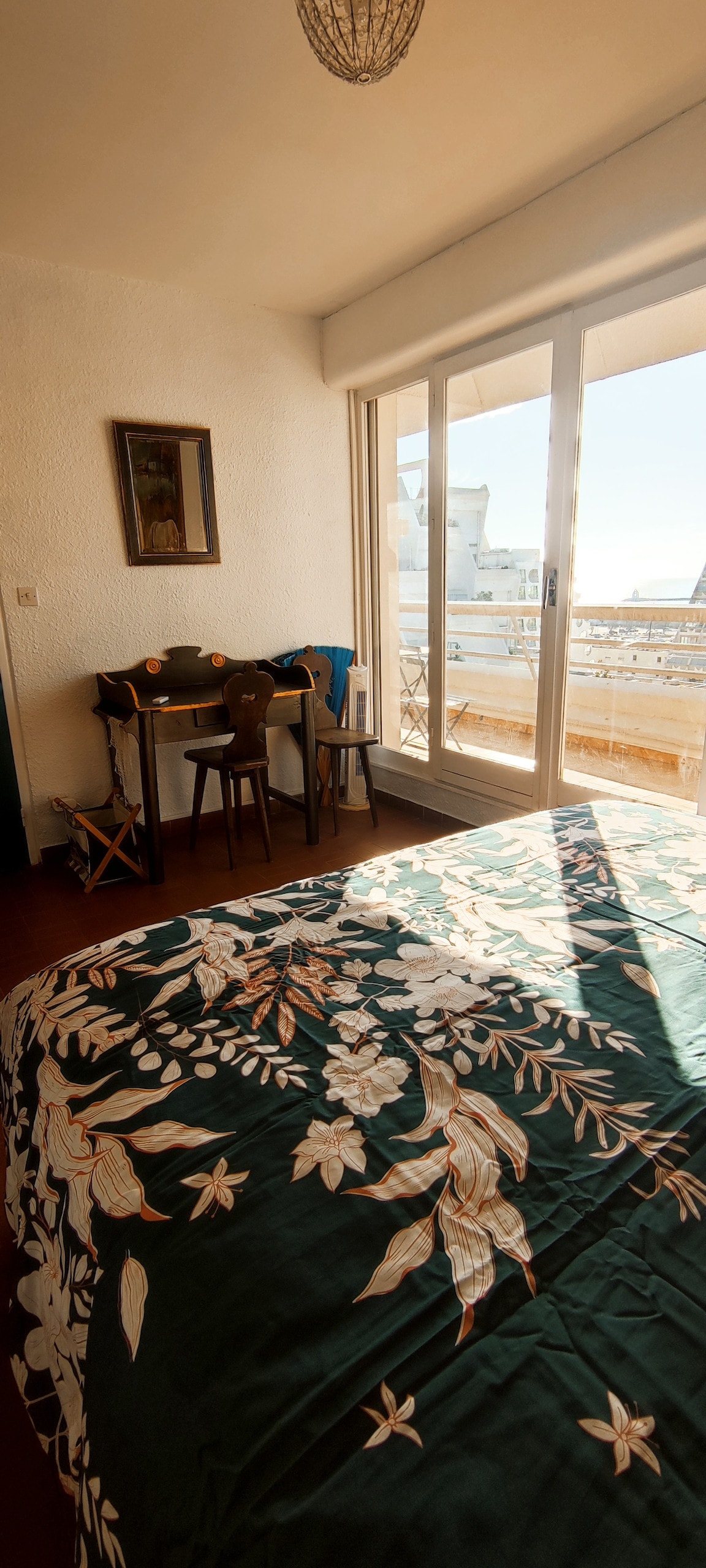 A well-lit bedroom features a spacious bed adorned with a green floral cover. A small table with two chairs is located beside a large window, offering a view of the surrounding area. Natural light floods the room, enhancing its open feel.