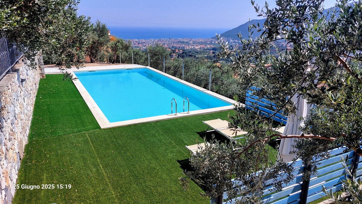 A serene outdoor space is featured, showcasing a clear swimming pool surrounded by lush greenery and well-maintained grass. Olive trees are visible nearby, with distant views of the coast and hills. Sun loungers are placed beside the pool, creating an inviting spot for relaxation.