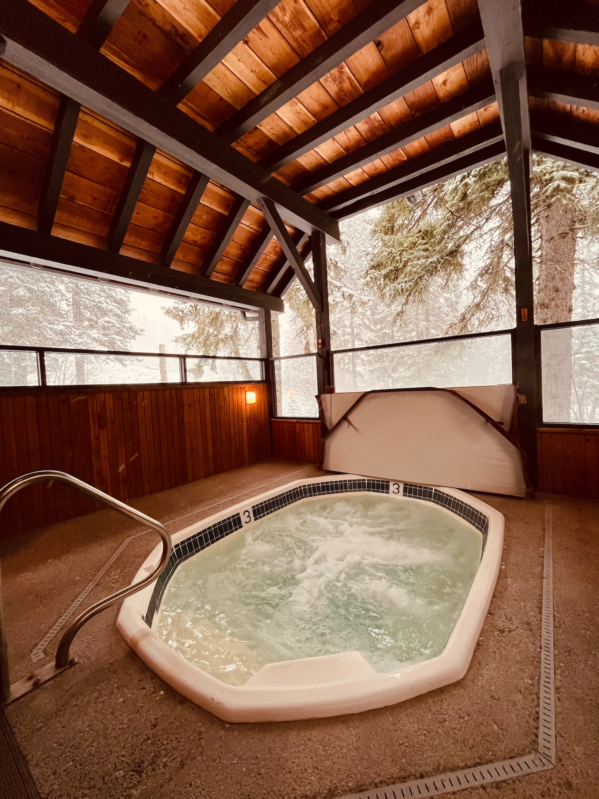 A hot tub is nestled within a cozy building featuring a high, wooden ceiling with exposed beams. Surrounding windows offer views of a snowy landscape outside, while soft lighting adds to the tranquil ambiance. A sturdy railing leads to the hot tub, enhancing accessibility.