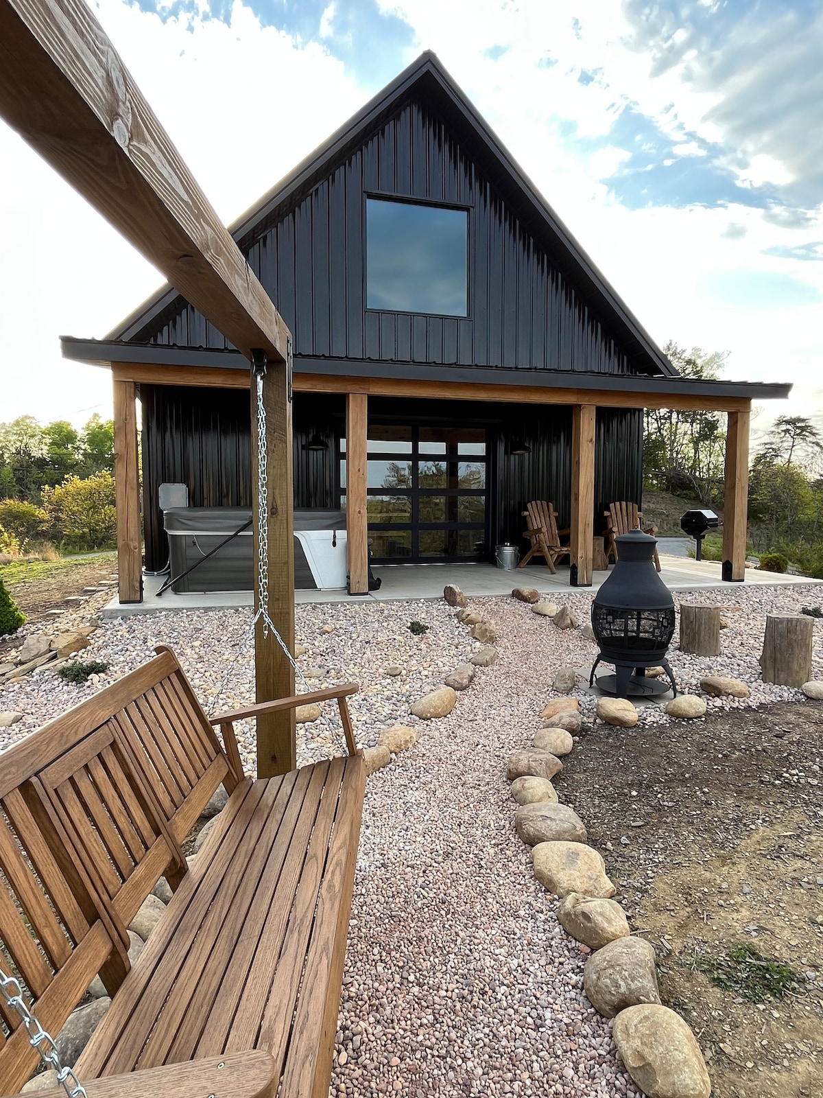 The cabin exterior features a striking black metal facade complemented by wooden beams and a large glass door. A swinging bench invites relaxation on the porch, while a fire pit surrounded by logs and stones enhances the outdoor space. Lush greenery is visible in the background.