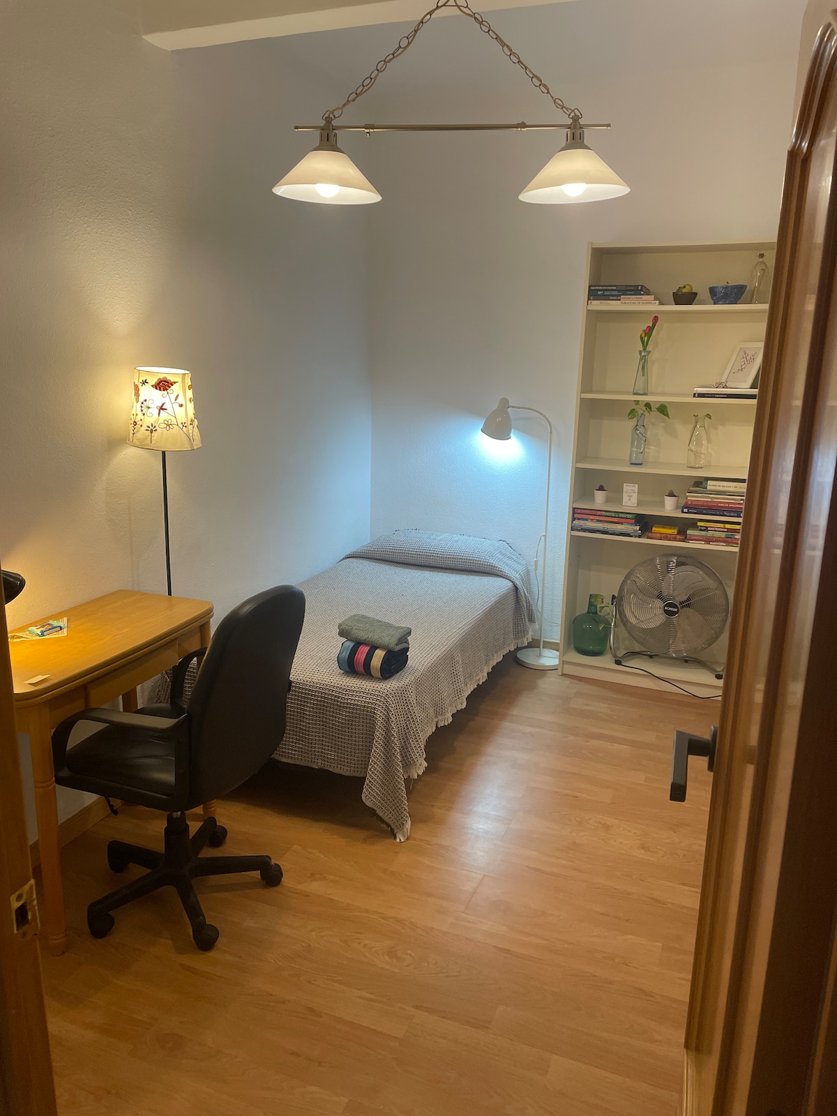 A simple bedroom is shown with a twin bed dressed in neutral linens. A small desk and rolling chair are positioned beside a light fixture. A bookshelf displays various items, and a standing lamp provides additional illumination. A fan is visible in the corner.