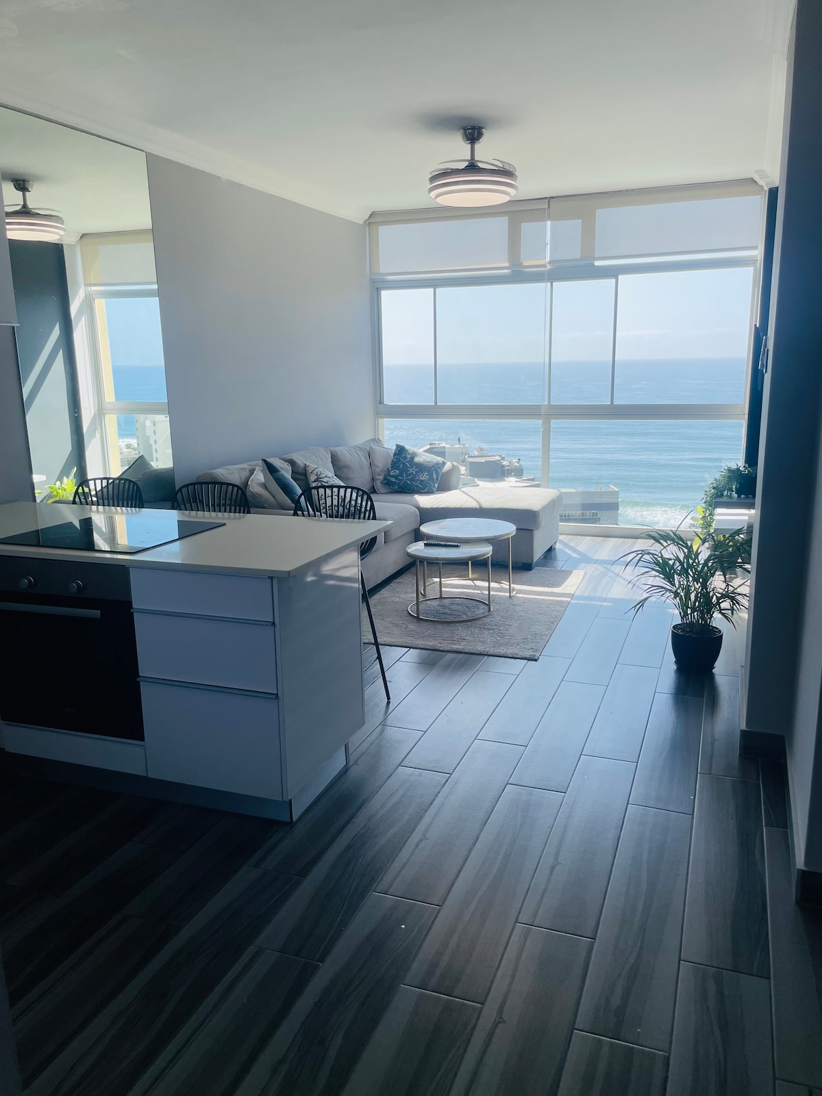 A bright living area is visible with a spacious sofa facing large windows that frame ocean views. A small round table sits on a rug, complemented by a decorative plant. The kitchen area features modern appliances, enhancing the open and airy atmosphere.