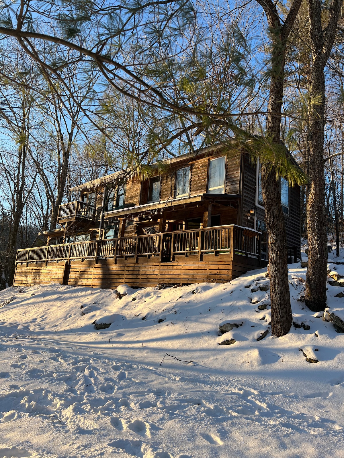 The wooden cabin is nestled among trees, with a snow-covered exterior that highlights its rustic charm. A spacious deck wraps around the front, inviting guests to enjoy the surroundings. Sunlight filters through the trees, creating a serene winter landscape.