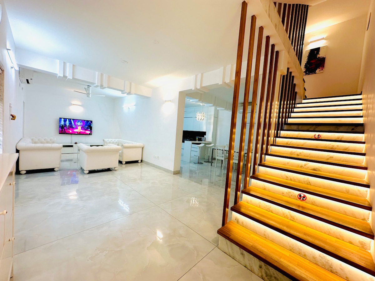 A light-filled living area features a white sectional sofa and a television mounted on the wall. A sleek staircase with illuminated steps leads to the upper level. The polished floor reflects the soft lighting, creating a spacious and inviting environment.