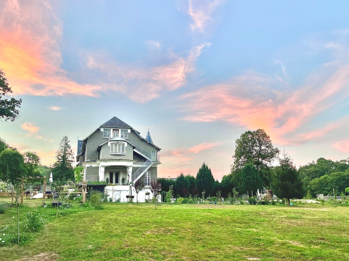 A spacious house with a multi-level facade is surrounded by lush greenery, sitting under a colorful sky at dusk. The property features a large lawn and various trees, creating a serene outdoor environment.