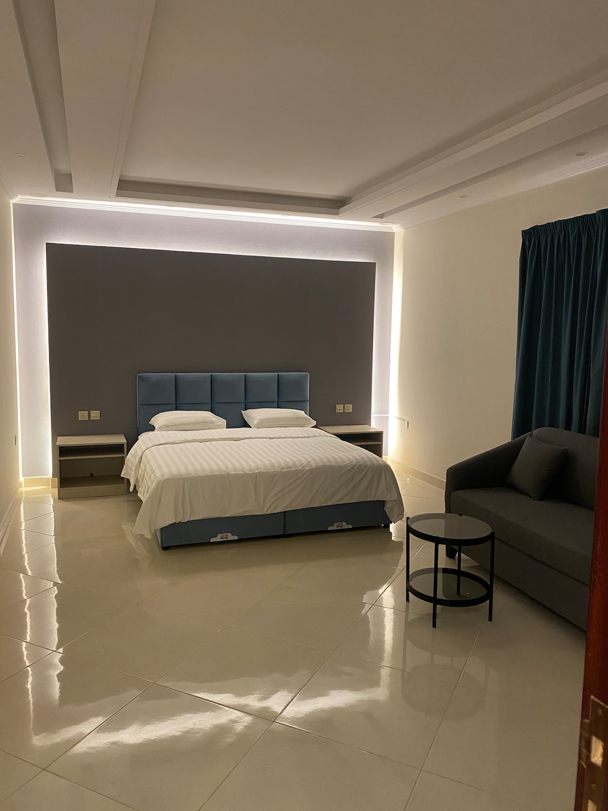 The bedroom features a spacious bed with a soft headboard, complemented by bedside tables on either side. A cozy seating area includes a small round table and a comfortable sofa. Soft lighting from the walls highlights the smooth tiled flooring and modern decor.