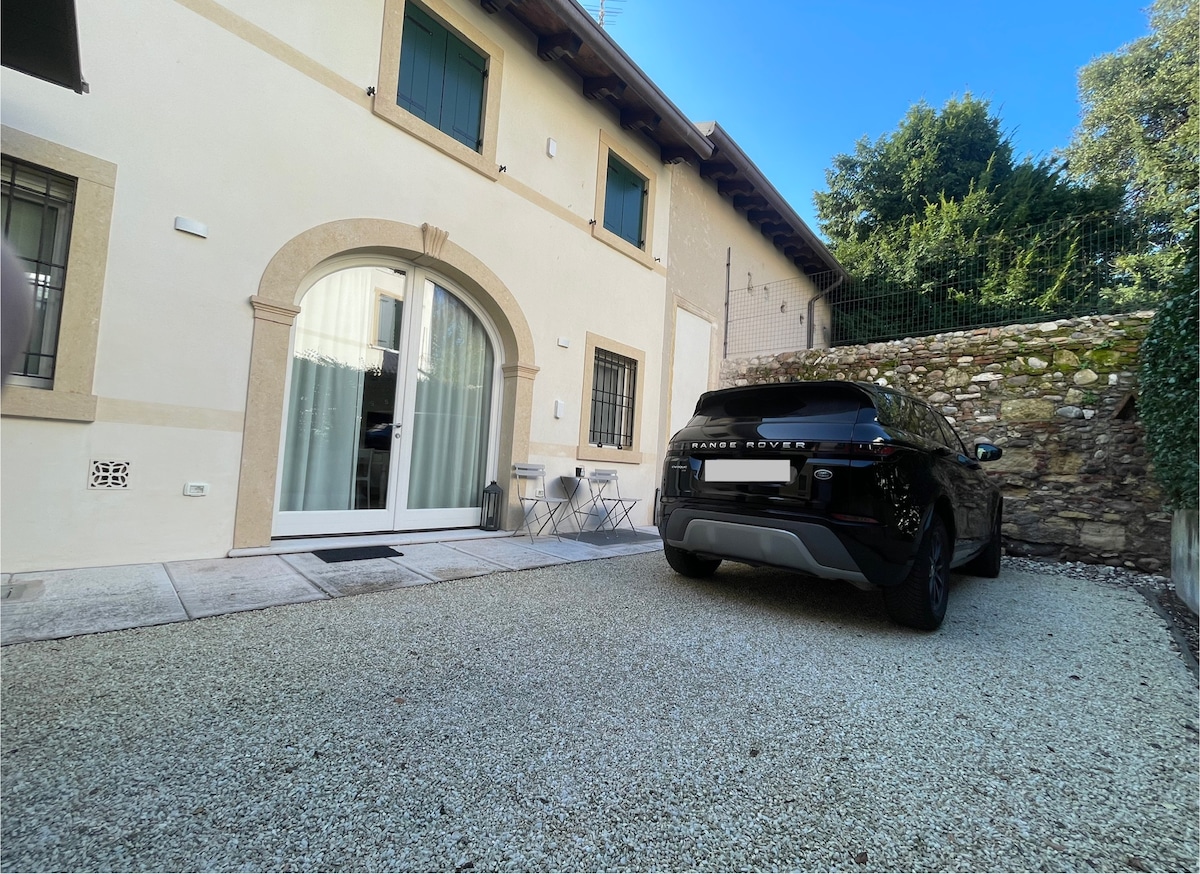 A private parking area is available next to the entrance, featuring a gravel surface. A modern vehicle is parked beside the building, which is characterized by a charming facade and large glass doors leading into the interior.