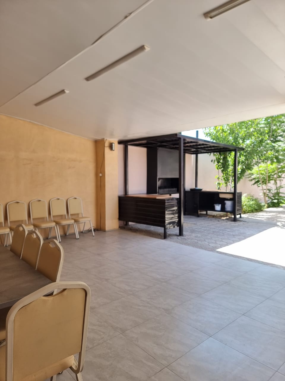 An open outdoor space features a covered seating area with several chairs arranged around a table. A barbecue area is positioned in the background, accompanied by greenery. Natural light illuminates the tile flooring, creating an inviting atmosphere for gatherings.