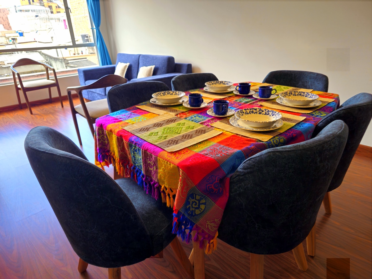 A dining table is set with colorful plates and mugs, surrounded by modern chairs. Nearby, a cozy living area features a blue sofa. Natural light filters through large windows, enhancing the warm ambiance of the space.