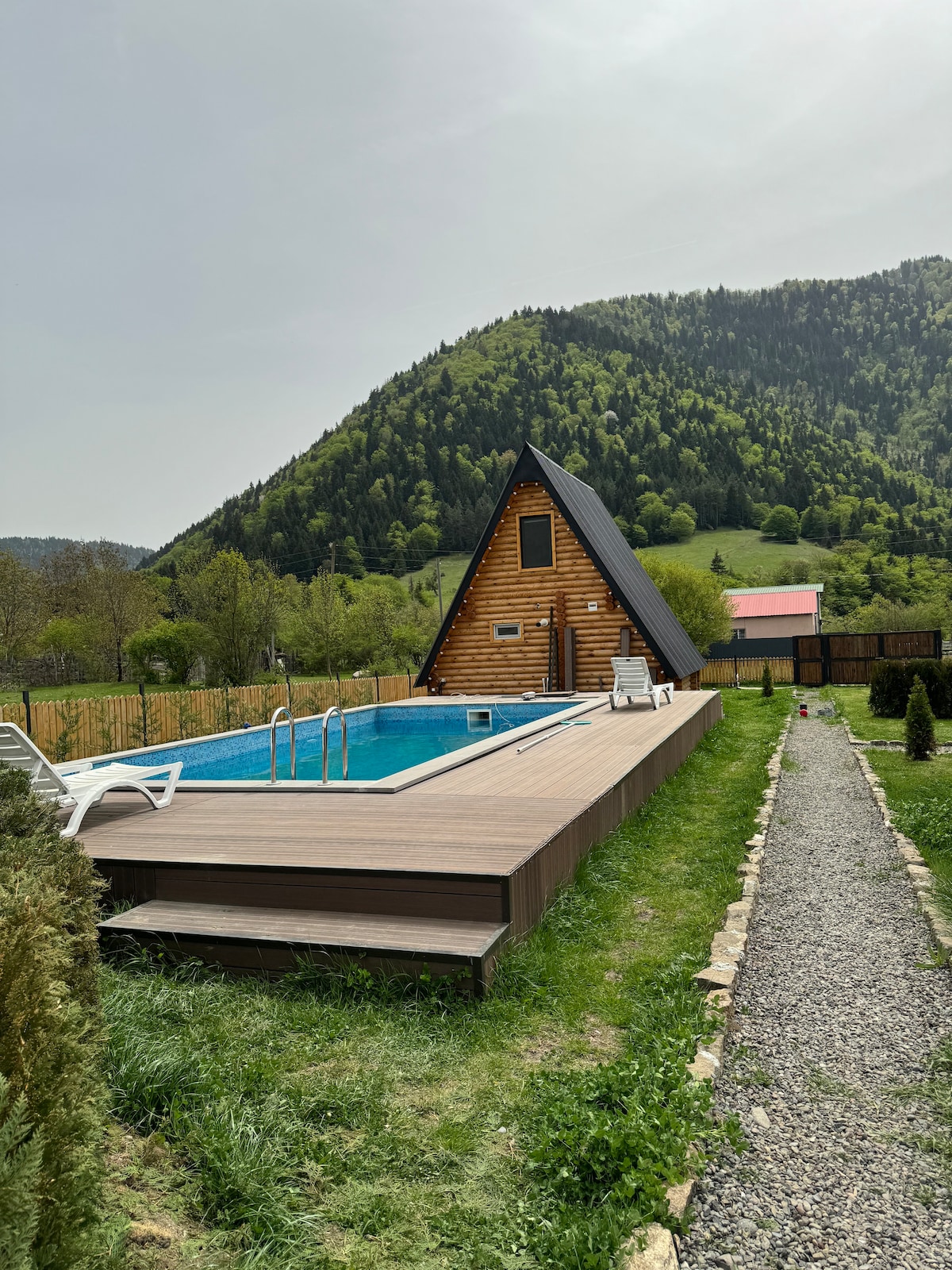 An A-frame cabin is situated near a clear blue pool, surrounded by a wooden deck. Lush green mountains rise in the background, complementing the serene landscape. A pathway of stones leads along the yard, flanked by manicured grass and shrubs.