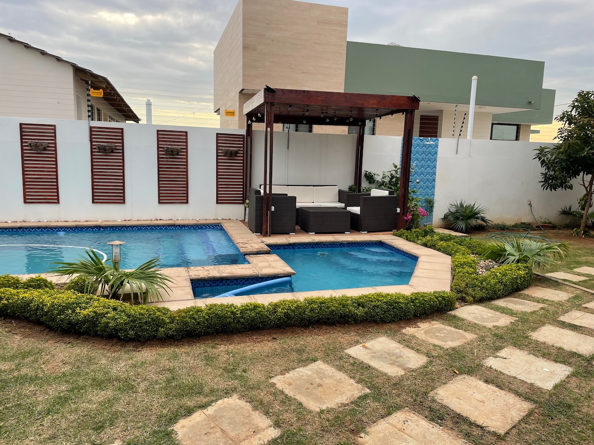 A landscaped outdoor area features a swimming pool surrounded by a stone patio. A shaded seating area is positioned beside the pool, with cushioned furniture and decorative plants enhancing the space. The neatly arranged garden includes a path made of stepping stones.
