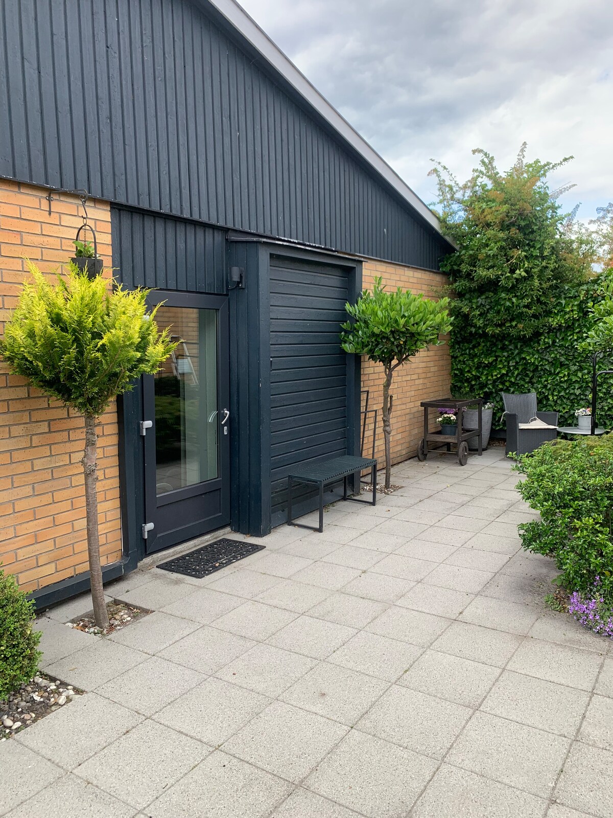 An outdoor area is presented, featuring a paved patio with a seating arrangement. Two small trees provide greenery alongside the space. A door with a glass panel leads into the building, and a small rug is positioned at the entrance.