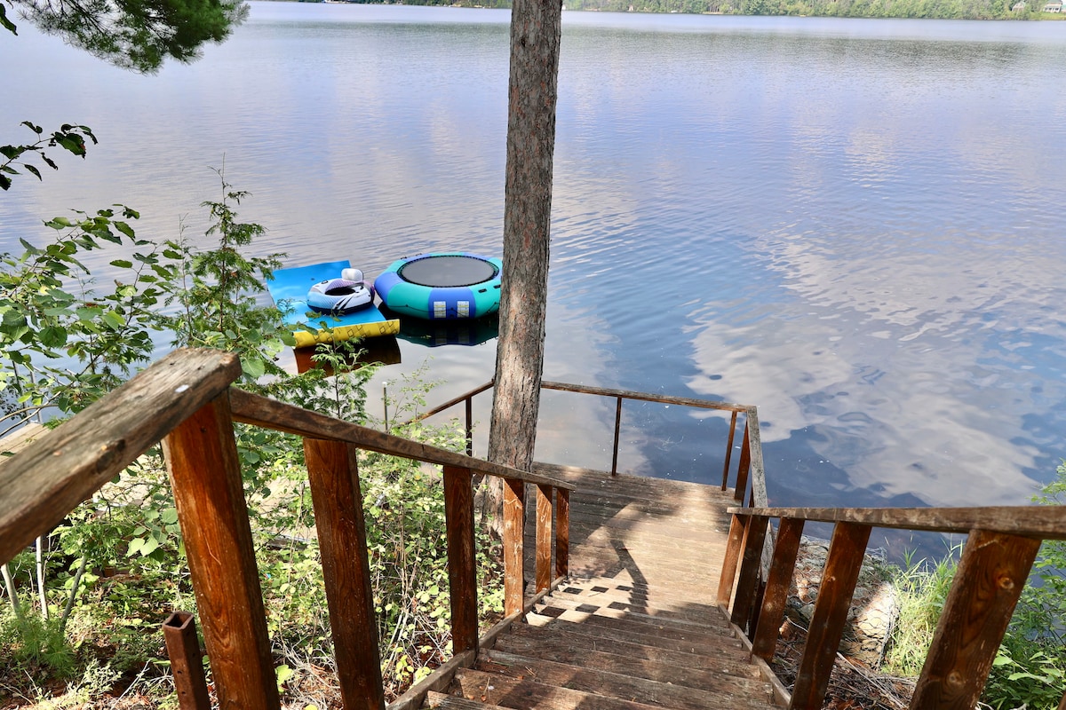 Sturdy wooden stairs descend toward a calm body of water, leading to a buoyant aquatic structure. Lush greenery frames the scene, enhancing the connection to nature. The surface of the water reflects the sky, contributing to a serene lakeside atmosphere.