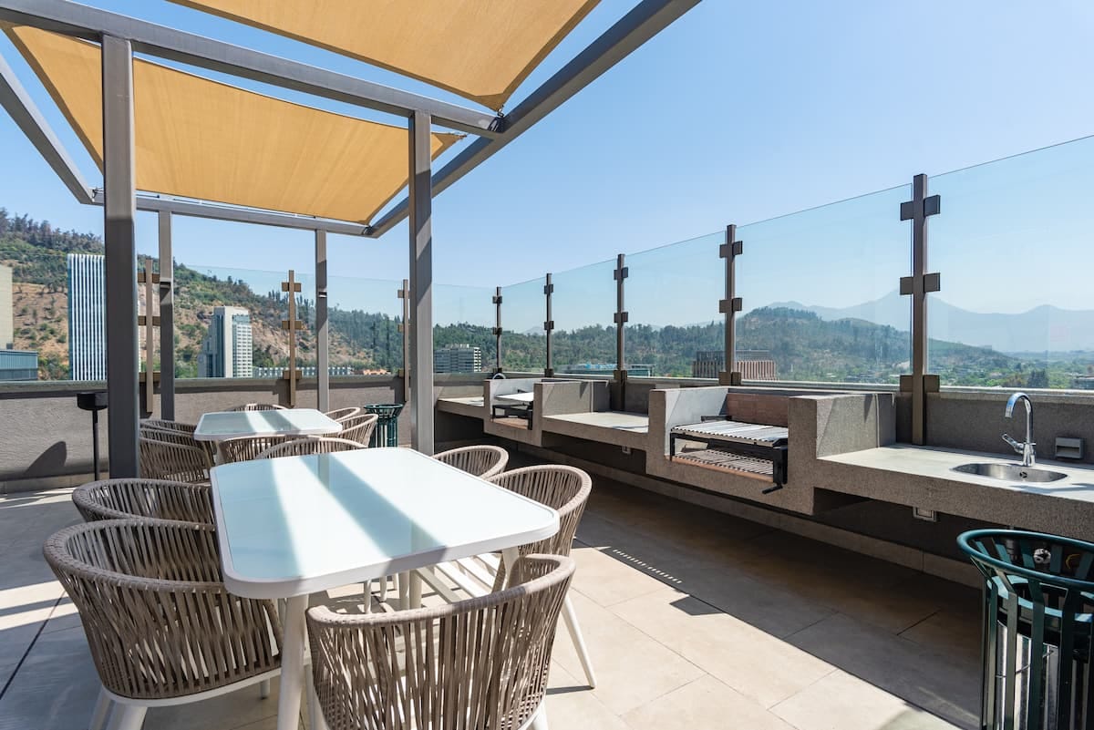A spacious rooftop terrace is equipped with several outdoor dining tables and chairs, offering sweeping views of the surrounding mountains and cityscape. Sun shades provide relief from direct sunlight, while a built-in grill and seating area invite social gatherings.